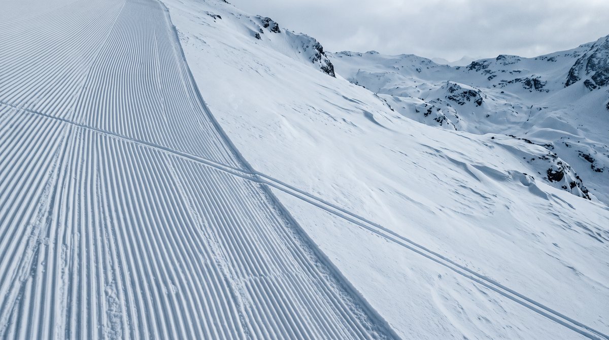 Le bord d’une piste damée vu de haut : le damage s’arrête net, une trace de skis le franchit vers la neige vierge.
