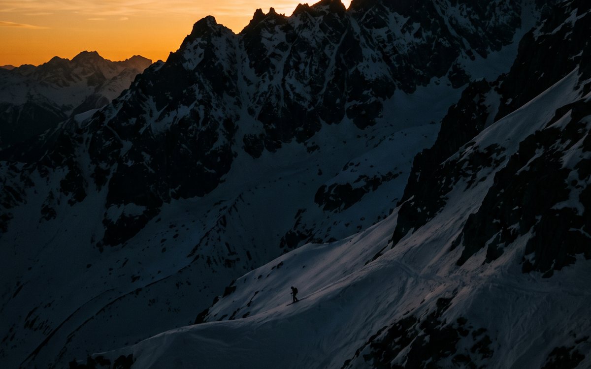 Une arête alpine en contre-jour au couchant, un skieur minuscule en silhouette sur la pente.